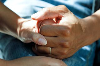 A close-up photo of two people holding hands. One person is wearing a wedding ring.