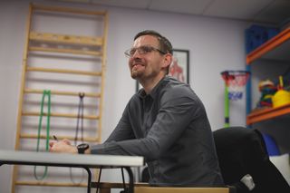 A man sat at a desk in the Second Chance rehabilitation centre. He is wearing glasses and smiling.